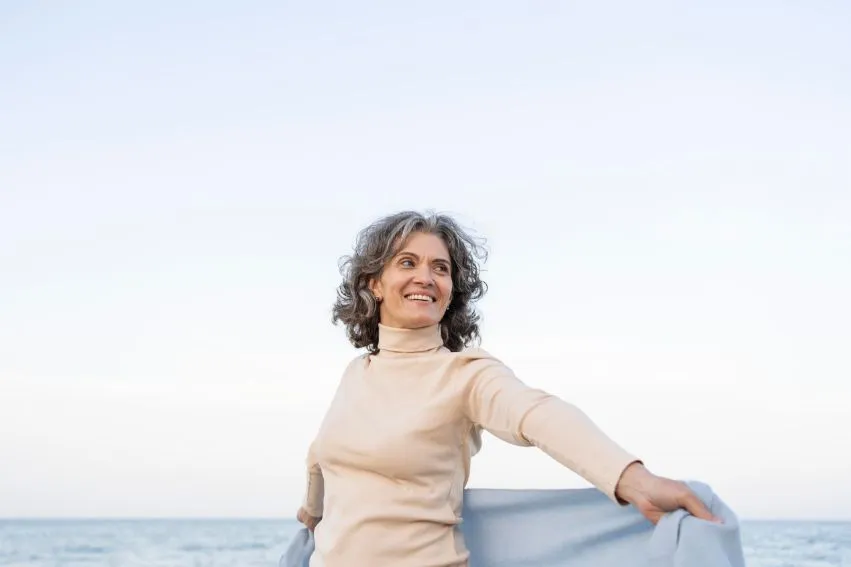 happy senior woman spreading her arms while standing on a beach, representing the benefits of Vagisil cream for vaginal health